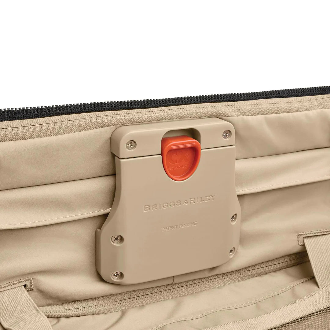 Close-up of a black Briggs & Riley Baseline Essential 22 Carry-On Expandable Spinner with an orange latch mechanism, logo, and PATENT PENDING. The CX expansion feature for added packing capacity is highlighted, alongside stitching detail and fabric texture.