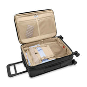 The Briggs & Riley Baseline Essential 22 Carry-On Expandable Spinner in black is shown open with a beige interior, containing a garment folder with neatly packed clothes, including a blue shirt and tie. It features compartments, two black wheels, and an extended handle for easy transport.