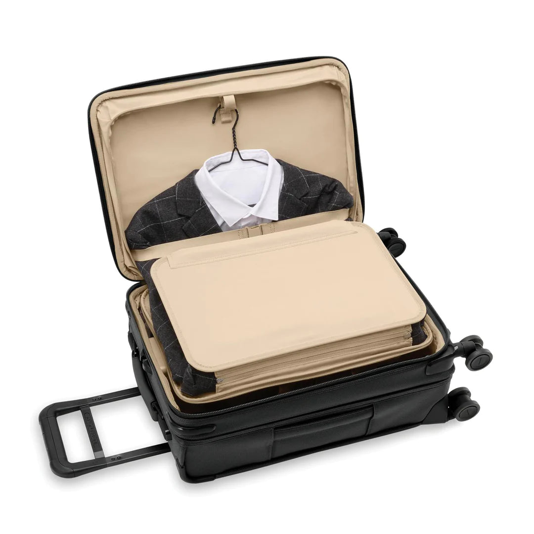 A black Briggs & Riley Baseline Essential 22 Carry-On Expandable Spinner reveals a gray checkered suit with a white shirt. The CX expansion adds space, while four wheels and a retractable handle ensure easy travel against a pristine white backdrop.