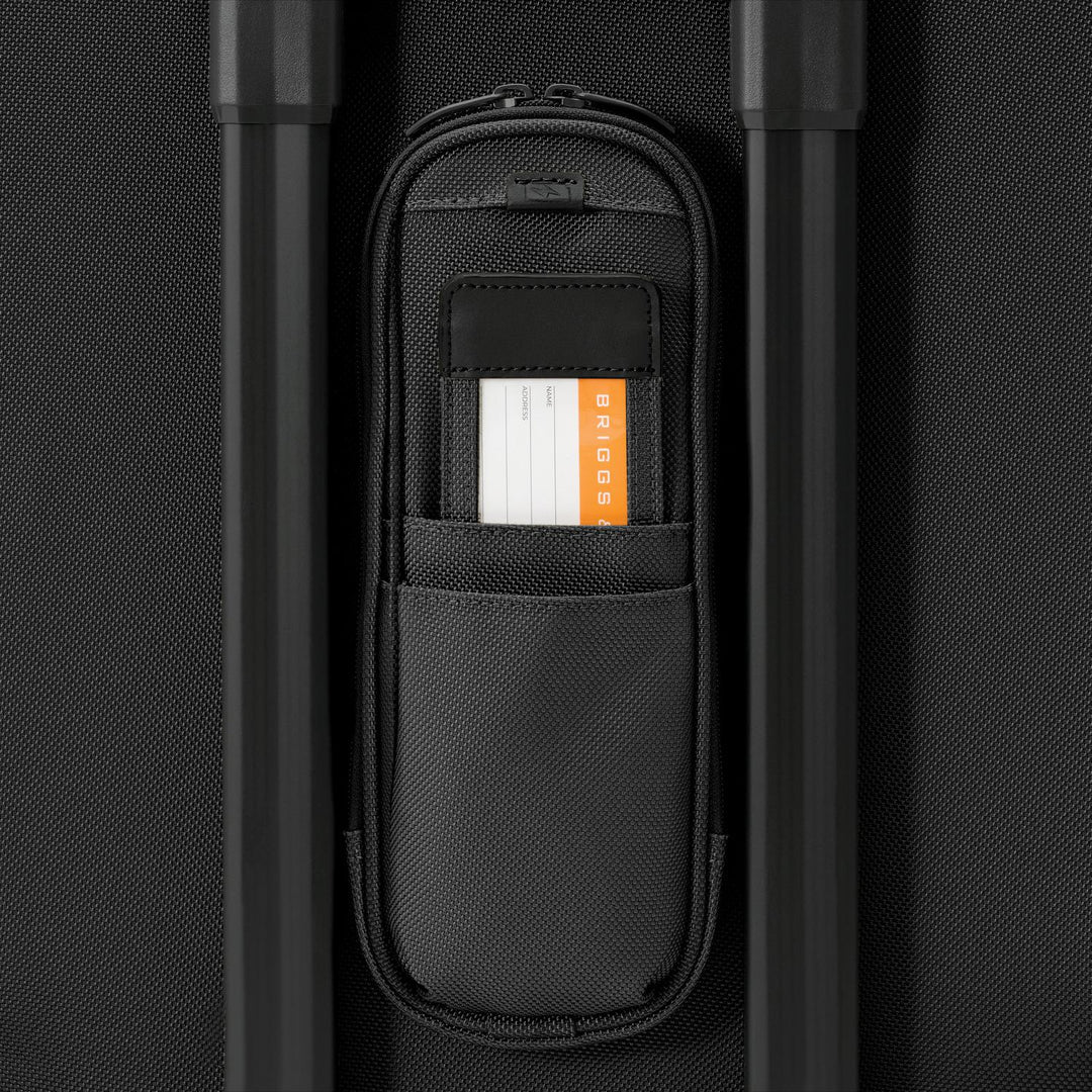 Close-up of a black Briggs & Riley Baseline Essential 22 Carry-On Expandable Spinner, featuring an open pocket with an BRIGGS ID tag between two handles on a textured black backdrop.