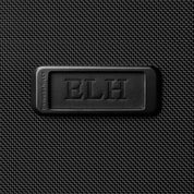 A black textured surface has a rectangular leather patch with ELH embossed in the center. The patchs corner reads BRIGGS & RILEY, indicating its part of the Briggs & Riley Baseline Essential 22 Carry-On Expandable Spinner - Black, featuring CX expansion capabilities.